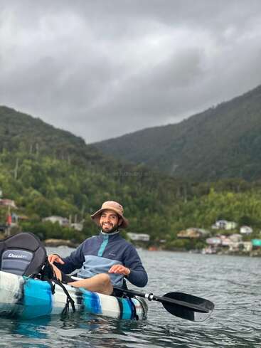 Um cara sorri enquanto anda de caiaque em um lago tranquilo, cercado por colinas verdes e casas espalhadas sob um céu nublado e encoberto. Ele usa um chapéu e uma jaqueta.