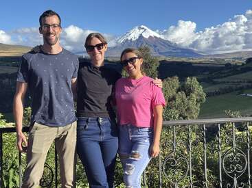 Drei Freunde lächeln zusammen auf einem Balkon, mit Sonnenbrille und lässiger Kleidung, mit einem schönen, schneebedeckten Berg und einer üppigen grünen Landschaft im Hintergrund.