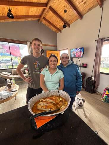 Drei Menschen posieren lächelnd in einem gemütlichen Haus mit Holzdecke. Ein Hund steht in der Nähe. Vorne auf dem Tresen steht ein großer Topf mit gekochtem Essen.