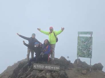 Four hikers celebrate atop a misty mountain summit, next to a sign reading "ALTITUD 4696 m s.n.m." and a post covered with colorful stickers.