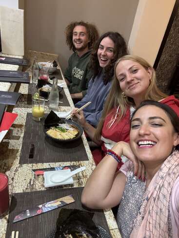 Four friends sit together at a restaurant, smiling and laughing. They enjoy Asian cuisine, using chopsticks, with drinks on the table, creating a joyful atmosphere.