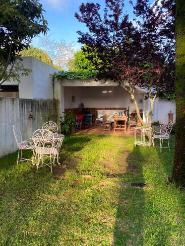 Un acogedor patio trasero con muebles de jardín metálicos, exuberante césped verde y árboles que dan sombra. Al fondo, un patio cubierto con mesas y sillas de madera invita a relajarse.