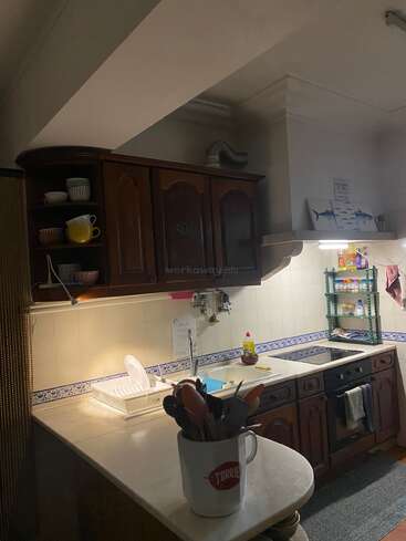 Essa cozinha aconchegante tem armários de madeira, fogão elétrico, vários utensílios em uma caneca, escorredor de pratos, prateleiras abertas e iluminação suave, criando um ambiente acolhedor e convidativo.