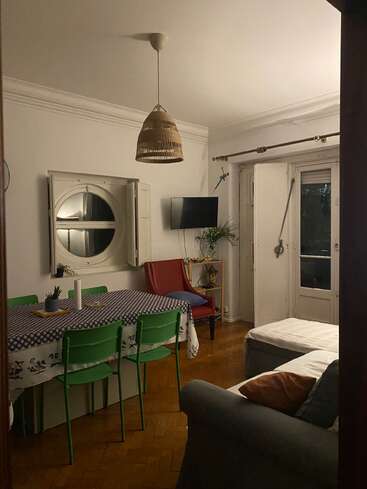 Uma sala de estar aconchegante com uma mesa grande, cadeiras verdes, uma poltrona vermelha, TV na parede, janela redonda, luminária pendente e portas de varanda. Piso de madeira em toda a sala.