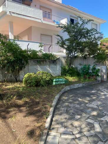Uma casa branca de dois andares com varandas e grades nas janelas. Tem vegetação ao redor, um caminho de pedras e uma placa verde de boas-vindas com os dizeres “Bem-Vindo Reception”.