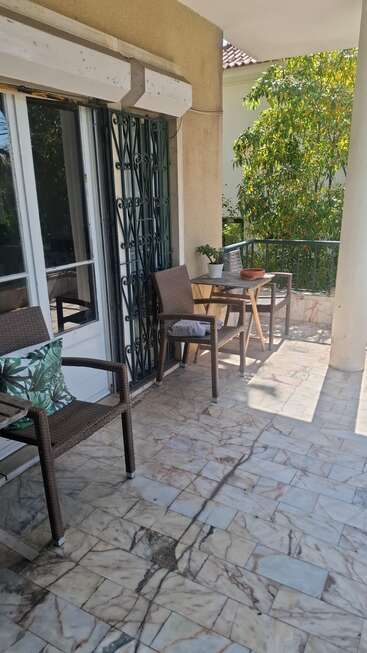 É uma varanda bem iluminada, com azulejos, cadeiras de vime, almofada, mesinha de madeira, vasos de plantas e vista para o jardim, perfeita pra relaxar ou tomar um café.