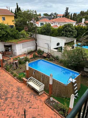 A imagem mostra um quintal com uma piscina acima do solo, pátio com azulejos, banco de mosaico, árvores, arbustos e casas ao redor com telhados de terracota sob um céu nublado.