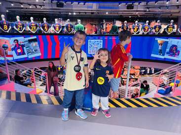 Three smiling children pose inside a vibrant football merchandise store, surrounded by colorful jerseys and digital screens. The atmosphere is lively, fun, and full of excitement.