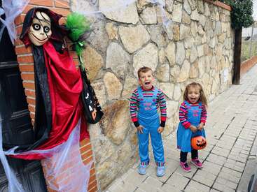 Deux enfants portant des costumes "Good Boy" assortis posent avec enthousiasme près d'un mur de pierre décoré pour Halloween, avec un personnage effrayant et des toiles d'araignée, en tenant des seaux de citrouilles.