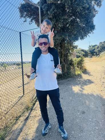 Un homme souriant porte un enfant heureux sur ses épaules en plein air. L'enfant fait des signes de paix. Ils profitent d'une journée ensoleillée près d'un grillage et d'arbres.