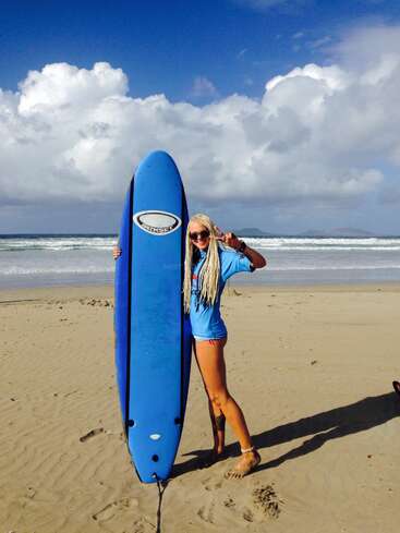 Uma mulher de cabelos loiros compridos está em uma praia de areia, segurando uma prancha de surfe azul, usando óculos escuros, sorrindo e fazendo o sinal da paz. Há ondas e nuvens ao fundo.