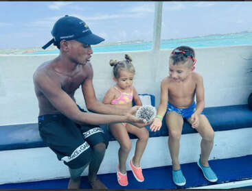 Um homem segura um ouriço-do-mar, mostrando-o a duas crianças pequenas de roupa de banho em um barco. As crianças parecem curiosas e animadas com a criatura marinha.