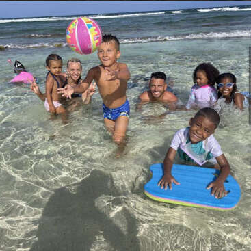 Eine Gruppe von Kindern und Erwachsenen spielt fröhlich im seichten Meerwasser. Ein Junge wirft einen bunten Strandball, während ein anderer ein blaues Boogie-Board in der Hand hält.