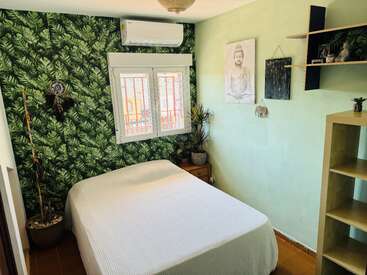 Este quarto aconchegante tem uma parede com detalhes em tons de verde, roupa de cama branca, ar-condicionado, janela, obra de arte com Buda, plantas, apanhador de sonhos, prateleiras de madeira e decoração em tons terrosos, criando uma atmosfera tranquila.