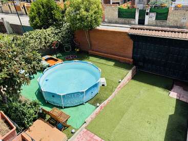 Esta imagem mostra um quintal com uma piscina acima do solo, grama artificial verde, um banco de madeira, plantas, árvores, equipamentos esportivos, um muro e uma garagem.