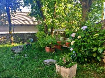 A imagem mostra uma cena de jardim sereno com vegetação exuberante, vasos de plantas e um banco, em uma parede de pedra rústica e um telhado de telhas.