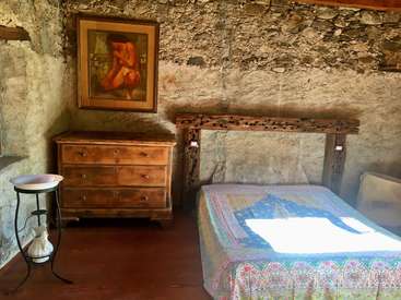 A imagem mostra um quarto rústico com uma parede de pedra, com uma cômoda de madeira, uma cama com uma colcha colorida e uma pintura de uma figura nua na parede.