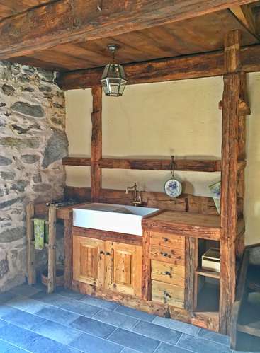 A imagem mostra uma cozinha rústica com pia e armários de madeira, com parede de pedra, teto de madeira e piso de ladrilho cinza, exalando um ambiente aconchegante e natural.