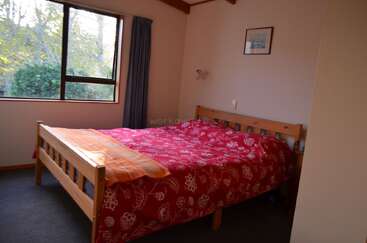 A cozy bedroom with a wooden bed frame, red floral bedspread, large window with a blue curtain, white walls, and a framed picture above the bed.