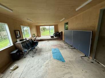 Une pièce spacieuse et inachevée avec un sol en béton, des chaises pliantes, un tapis bleu, une table de ping-pong, de grandes fenêtres, un mobilier basique et une vue sur un jardin.