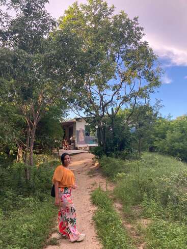 Eine Frau in einem farbenfrohen Outfit steht auf einem von üppigem Grün umgebenen Feldweg, der zu einem rustikalen Haus unter hohen Bäumen und blauem Himmel führt.