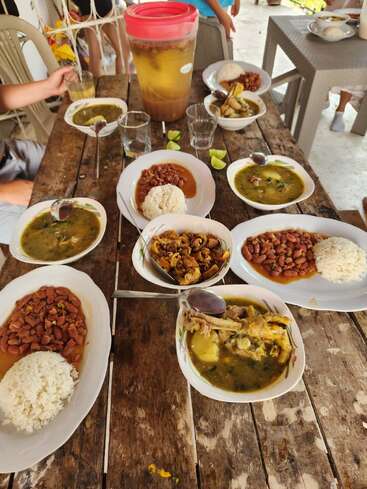 Uma mesa rústica de madeira está posta com pratos de arroz, feijão, sopa e curry, acompanhados por copos, fatias de limão e uma jarra grande de chá gelado.