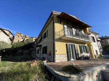 A two-story yellow house with green shutters, stone foundation, and a balcony. It has a spacious terrace, overgrown grass, and clear blue sky above.