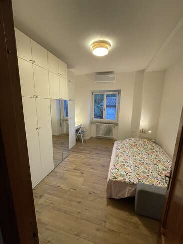 This image shows a tidy bedroom with wooden floors, a single bed with a floral cover, large white wardrobes, a desk, window, and ceiling light.