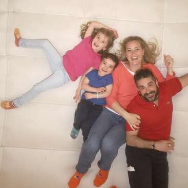 A happy family of four lies on a white surface. They are smiling and embracing, wearing colorful clothes. The moment captures warmth, love, and togetherness.