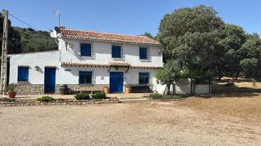 Une maison rustique blanche à deux étages, avec des portes et des fenêtres bleues, est entourée d'arbres sous un ciel clair, avec une cour en terre battue et un peu de verdure.