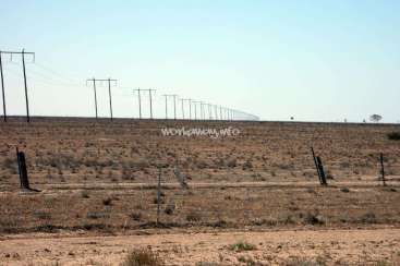 L'image représente un vaste paysage aride avec une rangée de lignes électriques s'étendant à l'horizon, sur fond de ciel bleu clair.