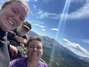 Tres personas sonrientes posan en la cima de una montaña rocosa, con impresionantes montañas, bosques y un cielo azul a sus espaldas. La luz del sol crea rayas brillantes sobre el fondo escénico.