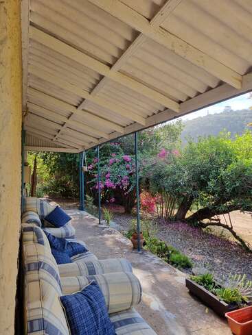 A cozy porch with cushioned chairs and blue pillows faces a beautiful garden with blooming pink flowers, lush greenery, and tranquil forested hills beyond.