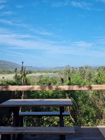 A wooden picnic table sits on a deck overlooking lush greenery and distant hills under a bright blue sky with scattered wispy clouds, creating tranquility.