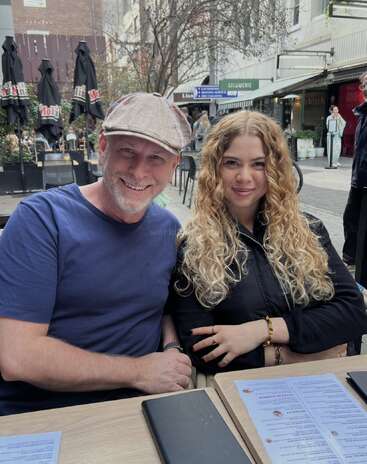 Un homme souriant portant une casquette et une femme aux cheveux bouclés sont assis ensemble à la table d'un café en plein air, les menus étalés, profitant d'une agréable journée.