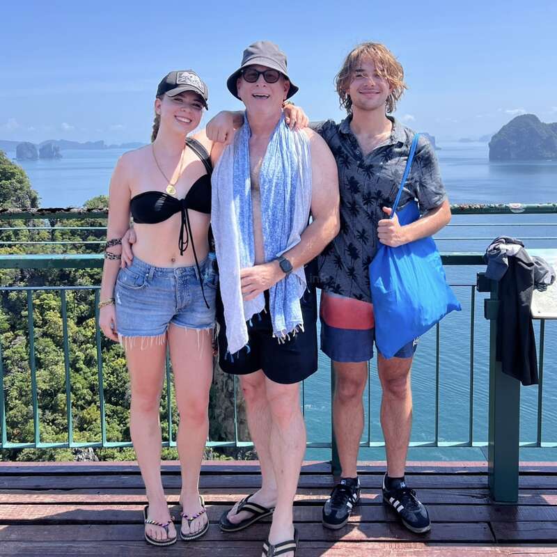 Trois personnes se tiennent ensemble sur un point de vue panoramique, en souriant. Derrière eux se trouvent de magnifiques eaux bleues, des îles et un ciel clair, suggérant des vacances tropicales ou une sortie en famille.