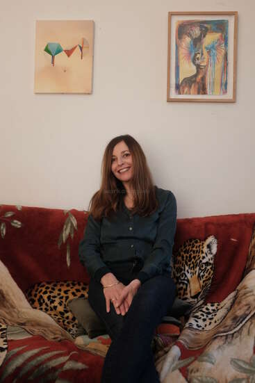 Eine Frau mit langen braunen Haaren sitzt lächelnd auf einem Sofa, das mit einer Decke mit Tigerprint bedeckt ist. Hinter ihr hängen zwei bunte Gemälde an einer schlichten Wand. Gemütliche Atmosphäre.