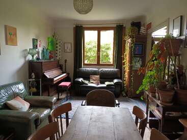 Ein gemütliches, wohnliches Wohnzimmer mit grünen Sofas, einem Klavier, Zimmerpflanzen, Holzmöbeln, bunten Gemälden und Tageslicht, das durch ein Fenster in der Mitte mit Vorhängen hereinfällt.