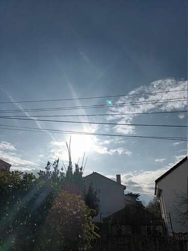 Das Bild zeigt eine strahlende Sonne, die hinter den Häusern scheint und einen Garten erhellt. Zu sehen sind einige Wolken, Stromleitungen, die über den Häusern verlaufen, und üppig grüne Büsche.
