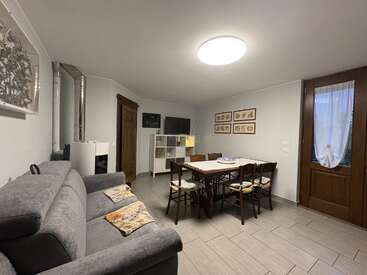 Esta imagen muestra un salón-comedor acogedor y amueblado con sencillez, con un sofá gris, una mesa de comedor de madera, sillas, estanterías, cuadros enmarcados y una puerta con cortinas de encaje.