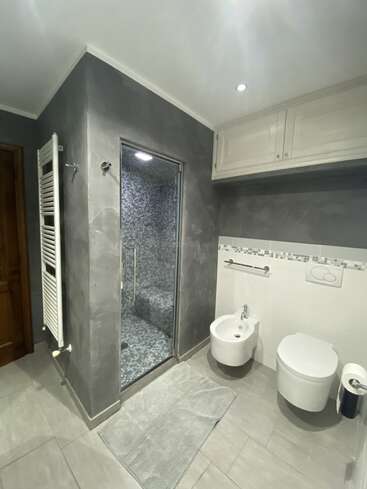 Este moderno cuarto de baño cuenta con una ducha acristalada con azulejos de mosaico, un inodoro suspendido, un bidé, muebles blancos, un calentador de toallas, paredes de color gris claro y una manta suave.