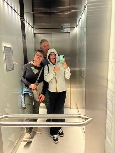 Tres personas posan para hacerse una divertida selfie frente al espejo dentro de un ascensor; una saca la lengua en tono juguetón, mientras que las otras se mantienen cerca, sonrientes y relajadas.