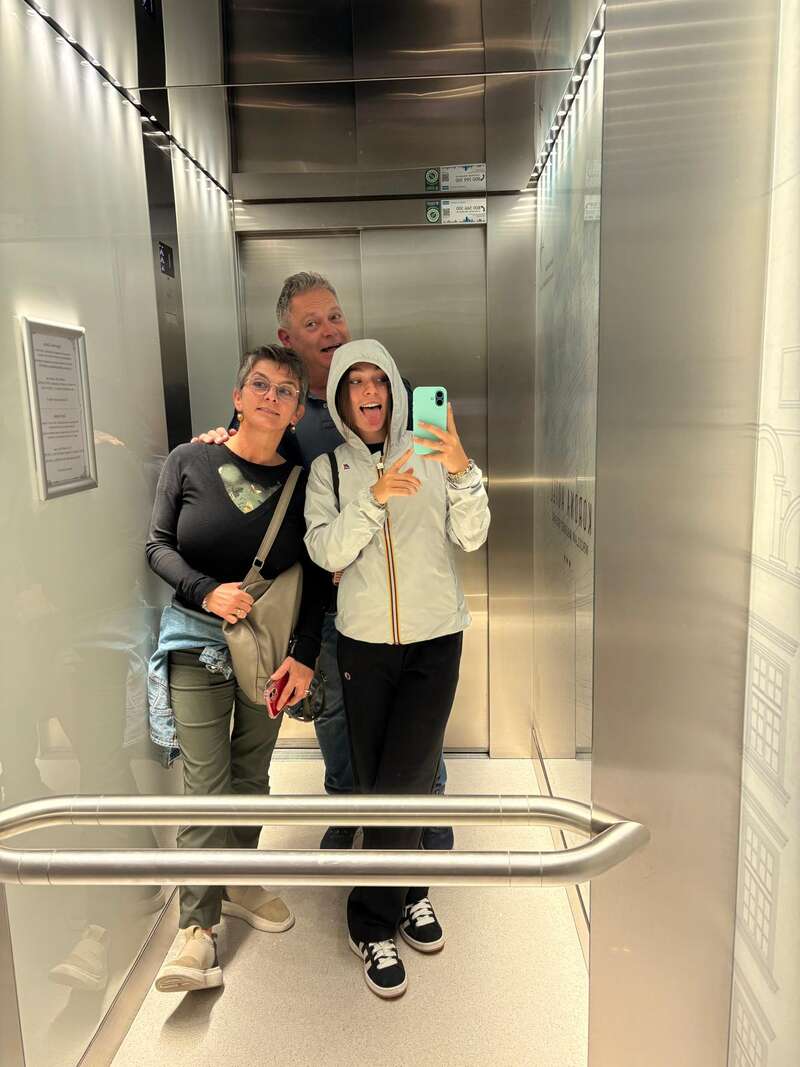 Tres personas posan para hacerse una divertida selfie frente al espejo dentro de un ascensor; una saca la lengua en tono juguetón, mientras que las otras se mantienen cerca, sonrientes y relajadas.