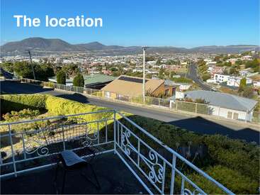 Esta imagen muestra un balcón con una silla de metal con vistas a un barrio residencial, al paisaje urbano y a montañas lejanas bajo un cielo azul despejado. Aparecen las palabras "La ubicación".