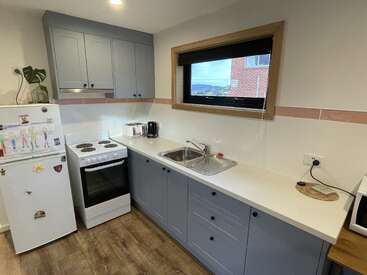 Esta cocina cuenta con armarios azules, encimeras blancas, un frigorífico con dibujos infantiles, cocina eléctrica, tostadora, hervidor de agua, fregadero, microondas, marco de ventana de madera y suelo de madera.