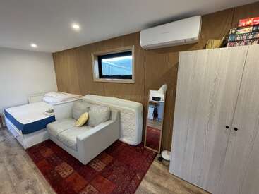 Esta acogedora habitación cuenta con una cama, un sofá con almohada amarilla, un armario, un aire acondicionado montado en la pared, un espejo y una ventana, todo ello sobre una vibrante alfombra estampada.