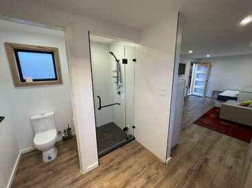 Esta imagen muestra un apartamento moderno con cuarto de baño con inodoro y ducha de cristal, suelo de madera, iluminación clara y una sala de estar contigua con sofá.