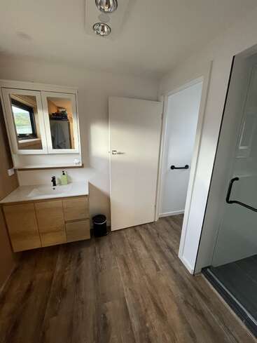 Este moderno cuarto de baño cuenta con suelo de imitación madera, un lavabo flotante con armario de espejo, una pequeña papelera, paredes blancas y una ducha con puerta de cristal y asideros negros.