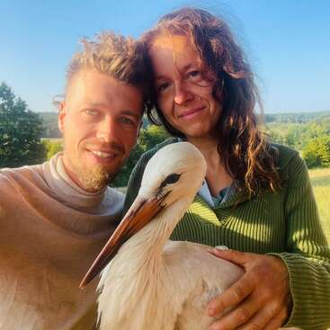 A smiling man and woman stand outdoors holding a white stork. The background features greenery, trees, and a clear blue sky, creating a serene, joyful scene.