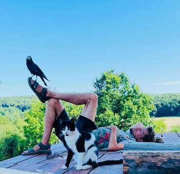 A man relaxes on a wooden deck in nature, lying down with a crow perched on his foot and a black-and-white cat sitting nearby, under clear skies.